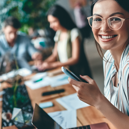 Smiling woman with a phone in her hand in front of a laptop. Other people and laptops in the background.