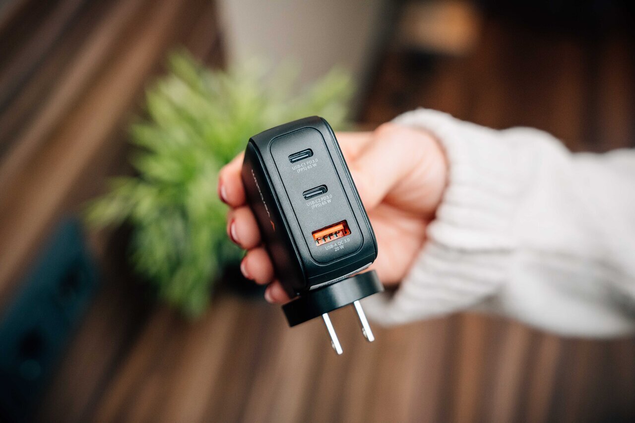 Person holding a GaN charger showing the USB-C and USB-A plugs on the end. Plant in the background.
