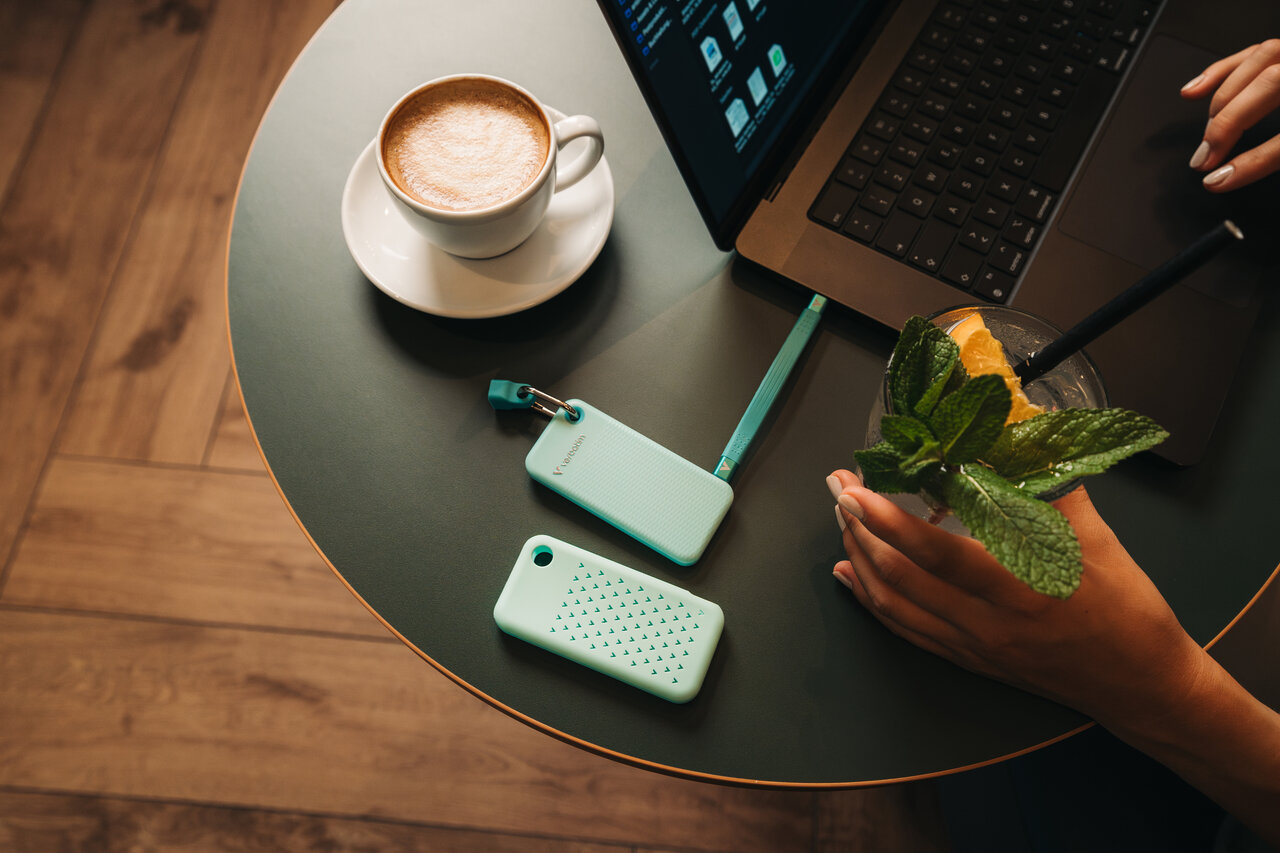 A arctic blue Verbatim Pocket SSD plugged into a laptop on a table in a coffee shop. A companied by a cup of coffee and a drink with mint garnish.