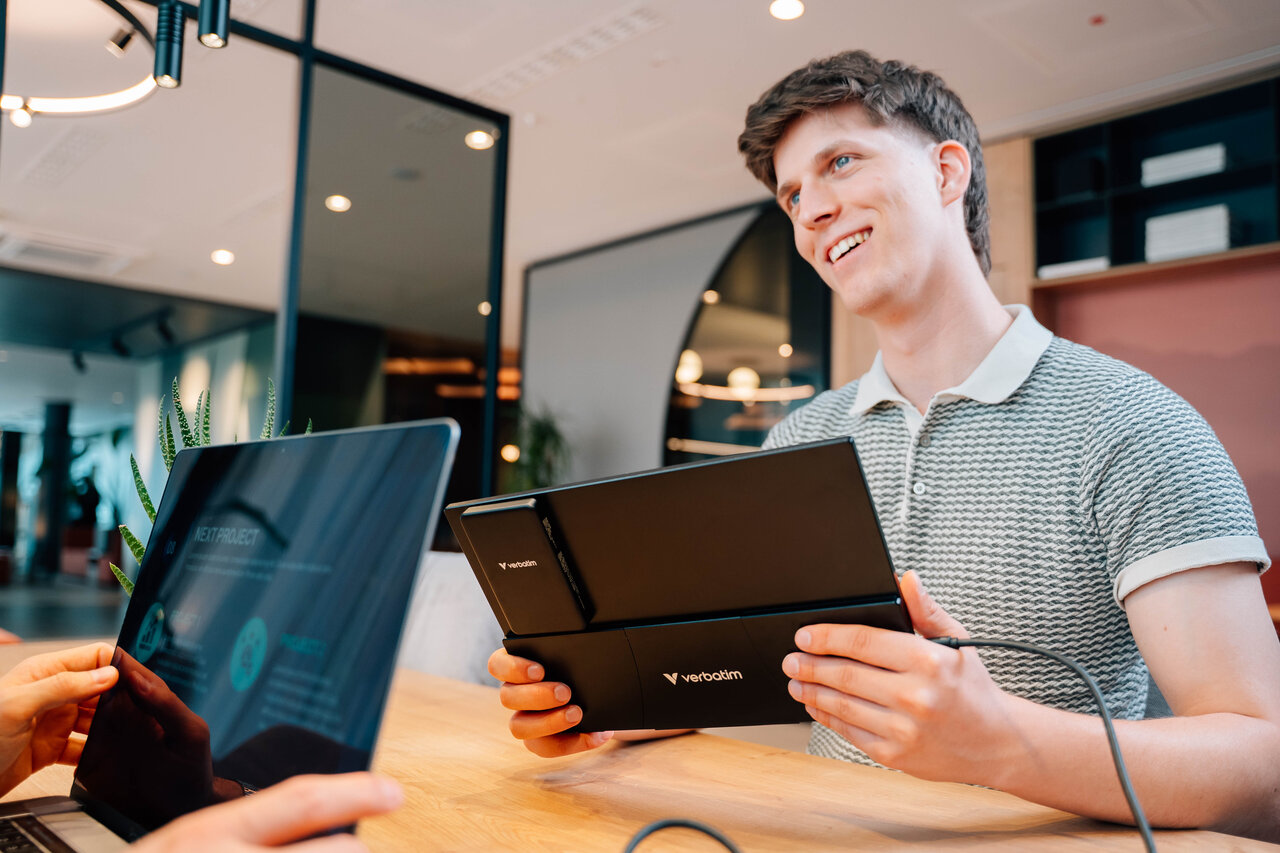 A man holding a black Verbatim portable monitor opposite another person with a laptop.
