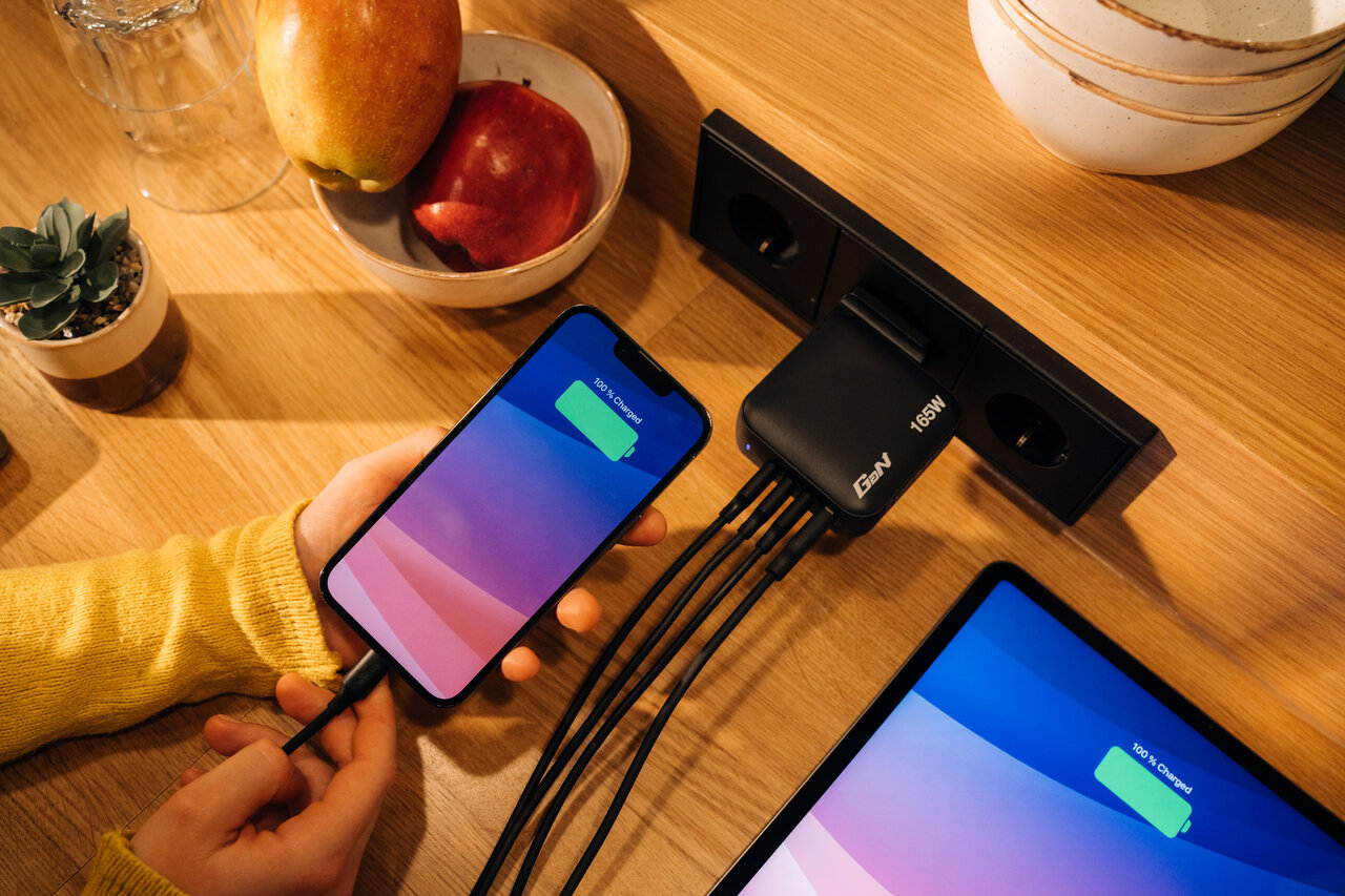 A GaN charger plugged into the wall, with 4 cables, charging an ipad and iphone. Fruit and plant on the desk nearby.