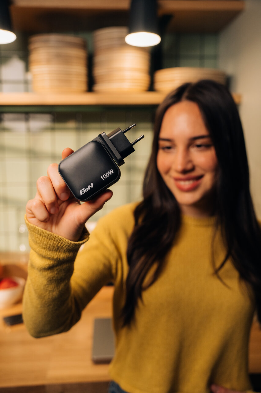 Woman with dark hair holding a 100W Gan charger with EU plug up. Kitchen in the background.