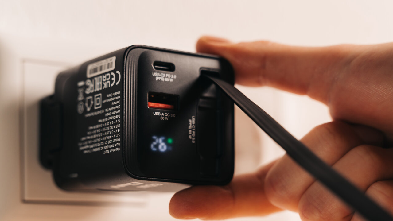 GaN charger being plugged into a wall socket with an integrated cable emerging from the back. 26 is shown on the digital display.