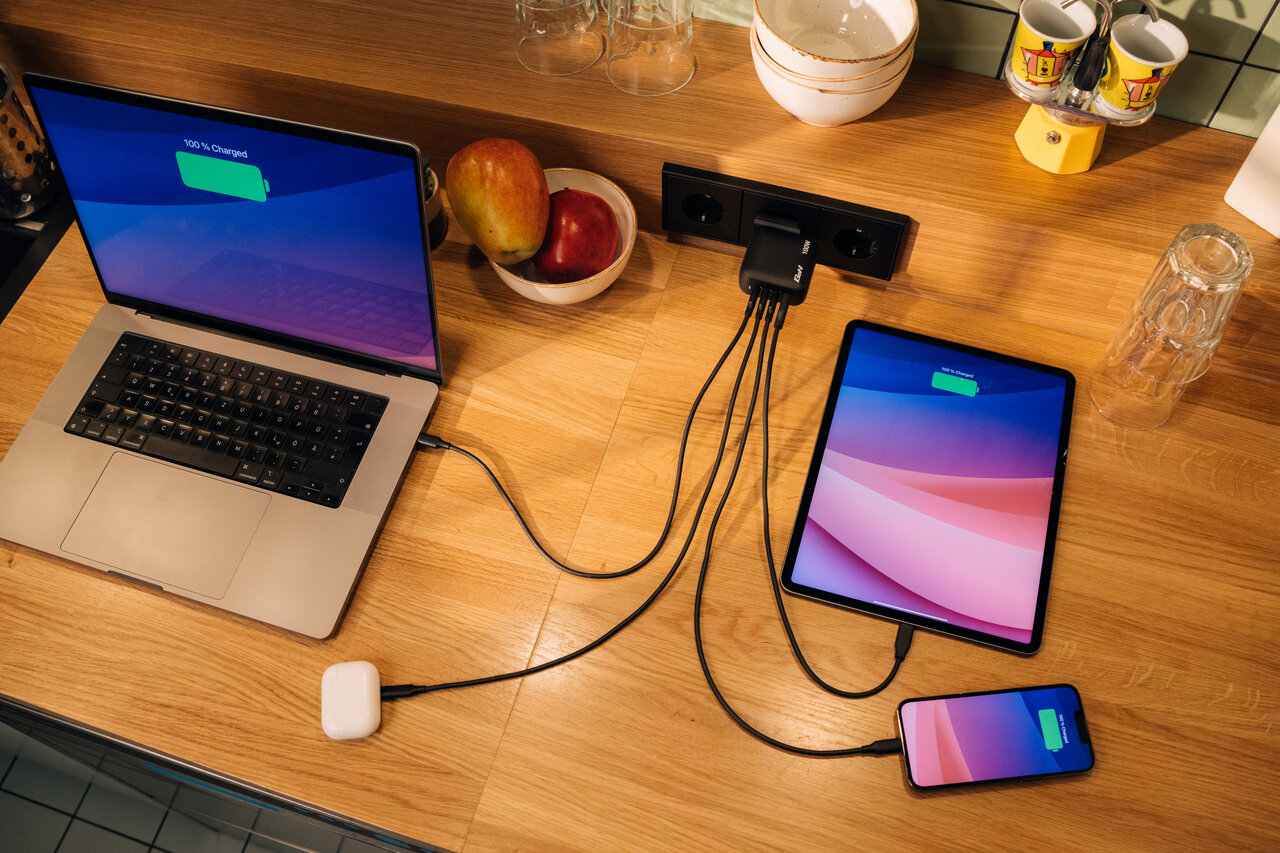Wooden desk with a laptop, AirPods, iPad and iPhone all plugged into a single charger plugged into the wall. All charged to maximum.